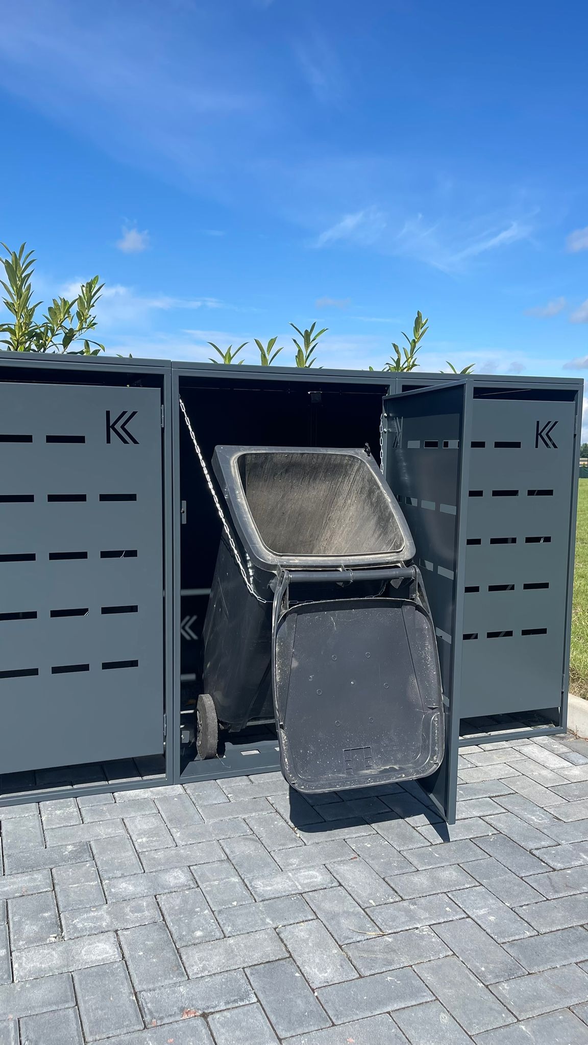 Metalowa obudowa na pojemniki na śmieci z ażurowym wzorem, umieszczona na kostce brukowej w słoneczny dzień, z otwartym pojemnikiem w środku.