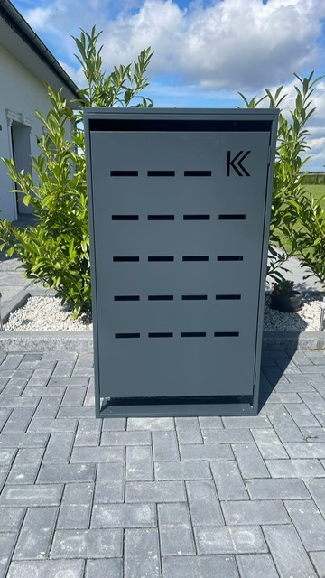 Szara, metalowa skrzynka na listy z poziomymi wycięciami i logo 'K' na tle kostki brukowej i zieleni.