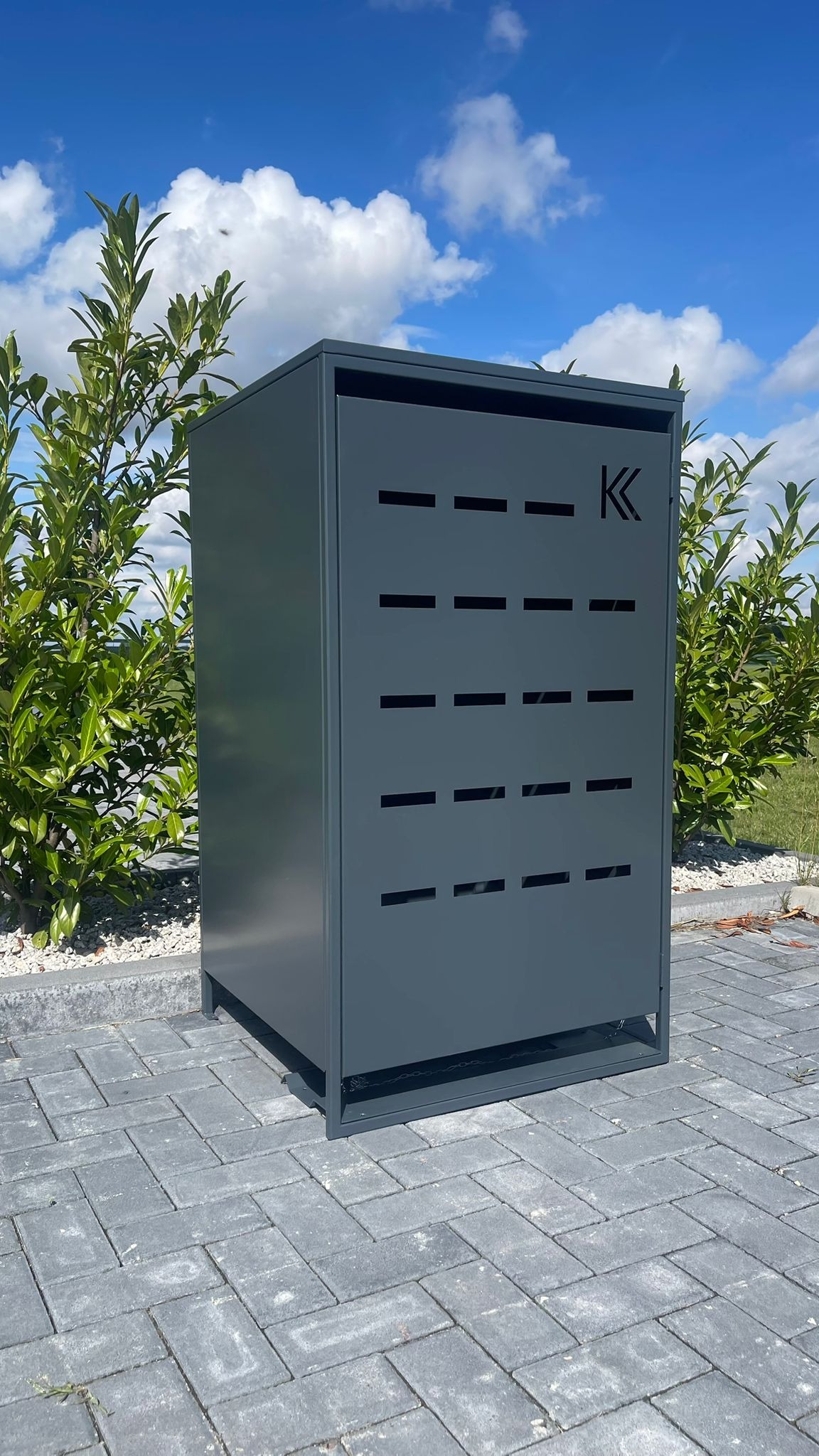 Szara szafka z metalu z poziomymi wycięciami i logo 'K' na tle niebieskiego nieba i zielonych krzewów, stojąca na szarej kostce brukowej.