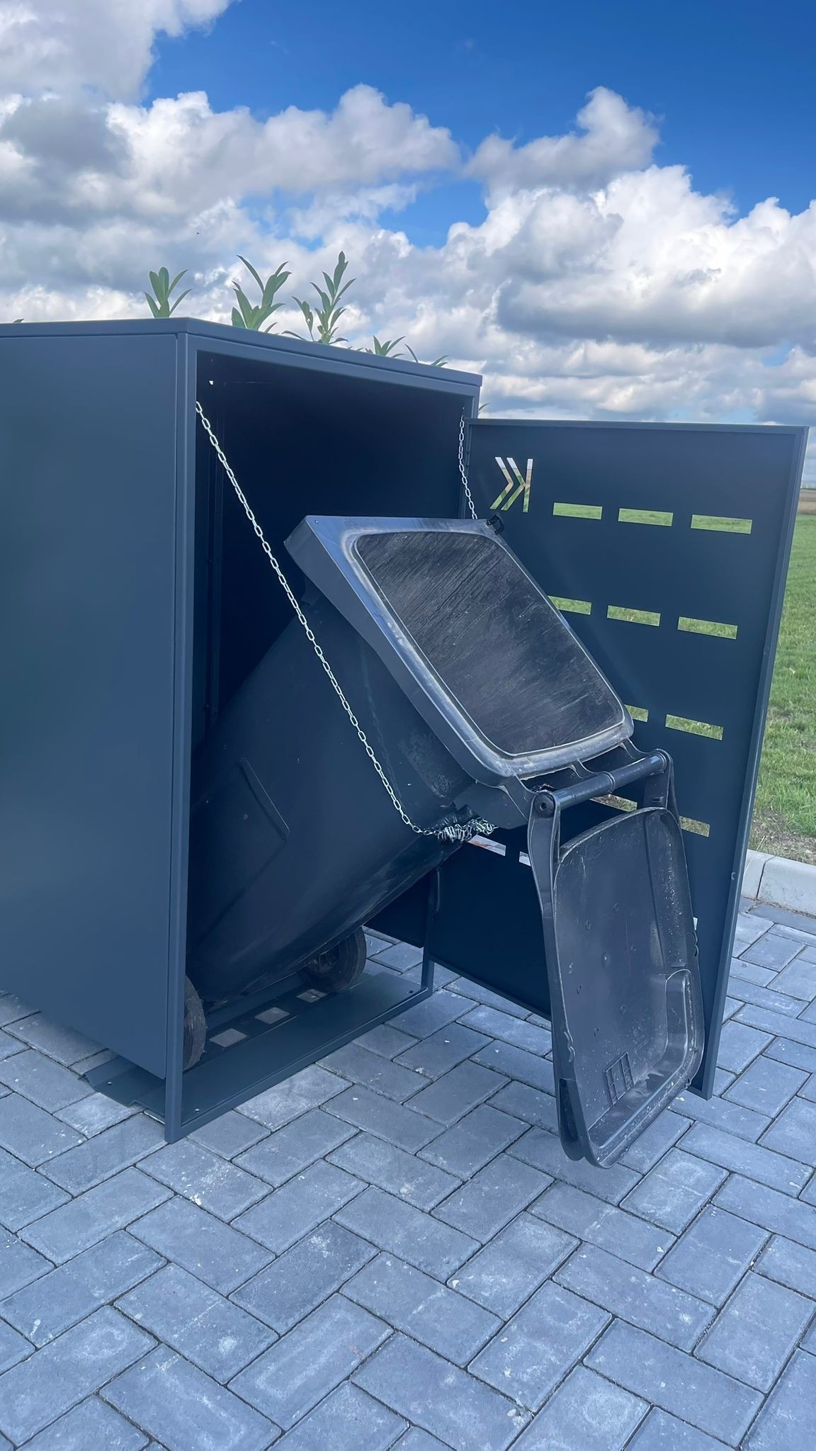 Metalowa obudowa śmietnika w kolorze grafitowym, z ażurowym wzorem i roślinami na dachu, umieszczona na brukowanej powierzchni.