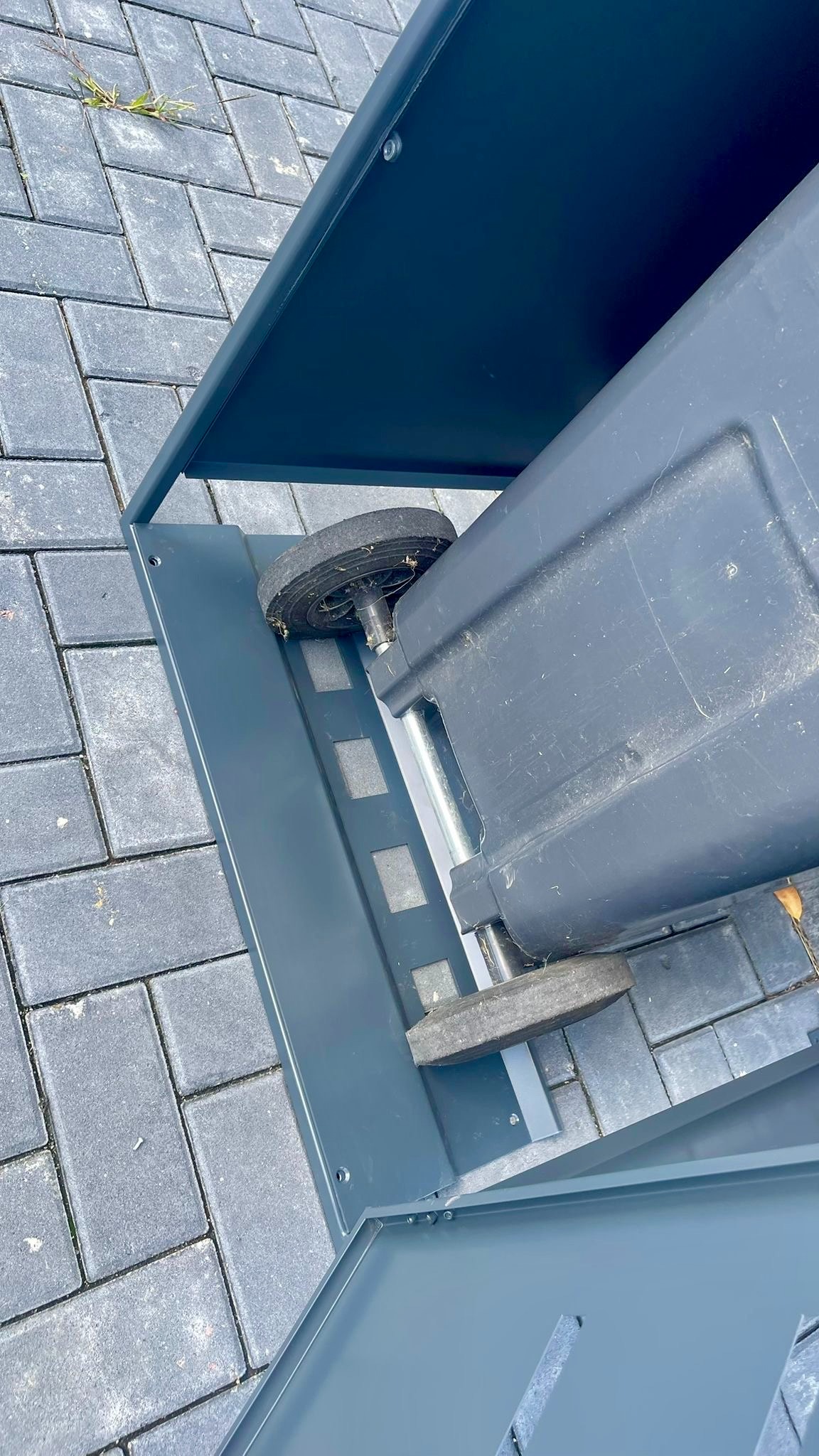 Metalowa obudowa na pojemnik na śmieci w odcieniach szarości, z widocznym kołem pojemnika, na tle szarej kostki brukowej. Widok z góry.