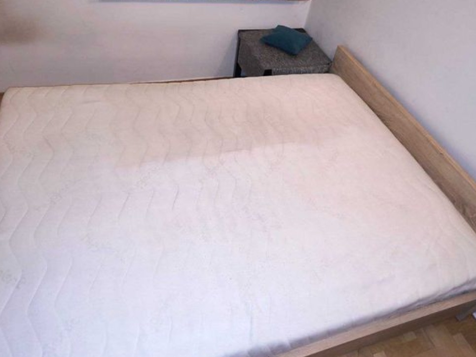 Wyprany materac na łóżku z drewnianą ramą, obok szafka nocna z poduszką. Widoczne ślady po praniu na powierzchni materaca.