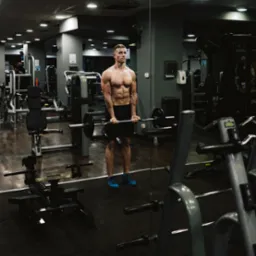 Mężczyzna w sportowych butach i spodenkach, bez koszulki, ćwiczy z obciążeniem na tle siłowni. Widoczne maszyny i lustro.