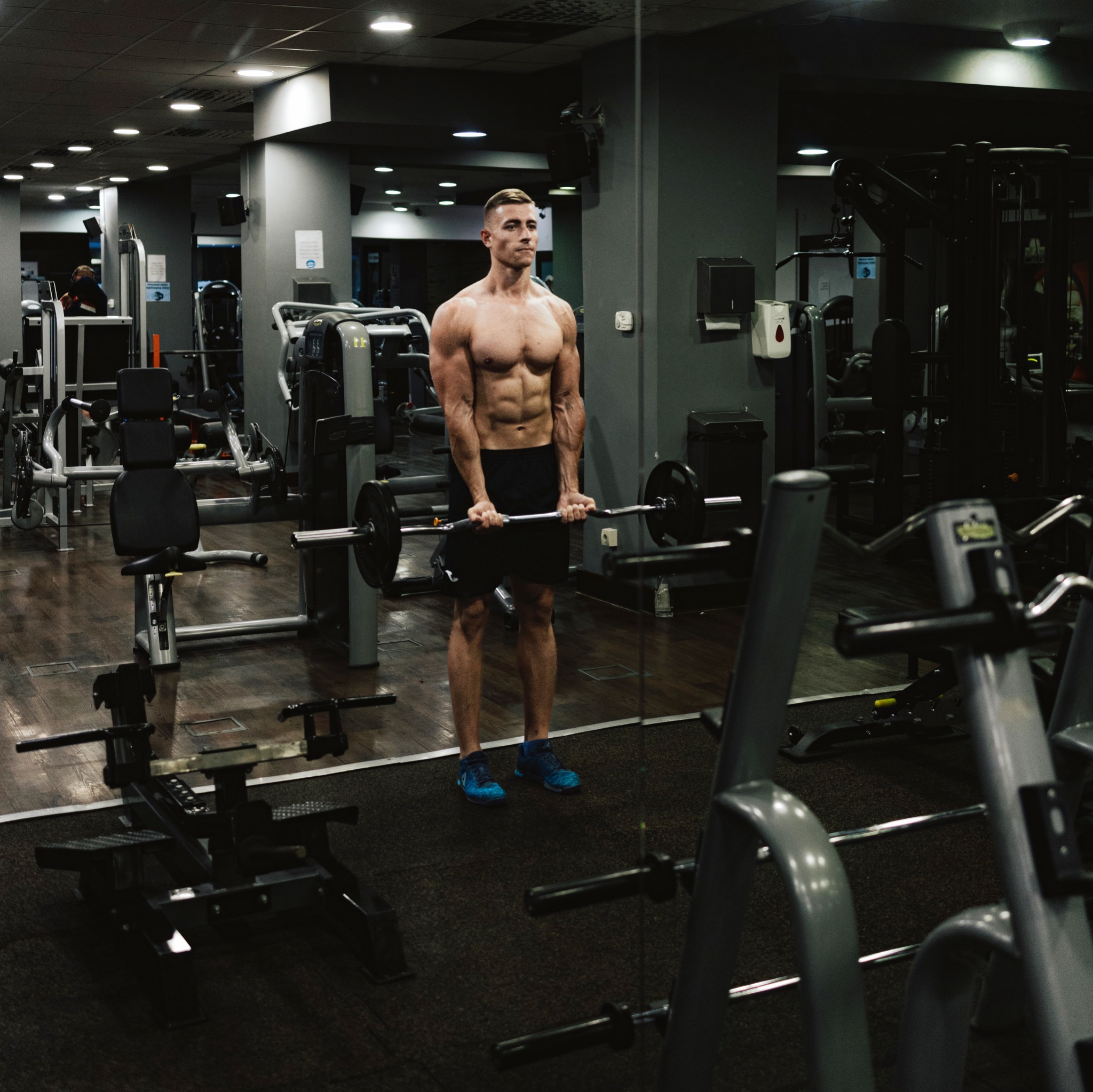 Mężczyzna w sportowych butach i spodenkach, bez koszulki, ćwiczy z obciążeniem na tle siłowni. Widoczne maszyny i lustro.