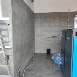 Krzysztof PYS - Szare, prostokątne płytki ścienne i podłogowe w pomieszczeniu z instalacją grzewczą. Widoczna drabina, rury i zbiornik na wodę. Minimalistyczny design.
