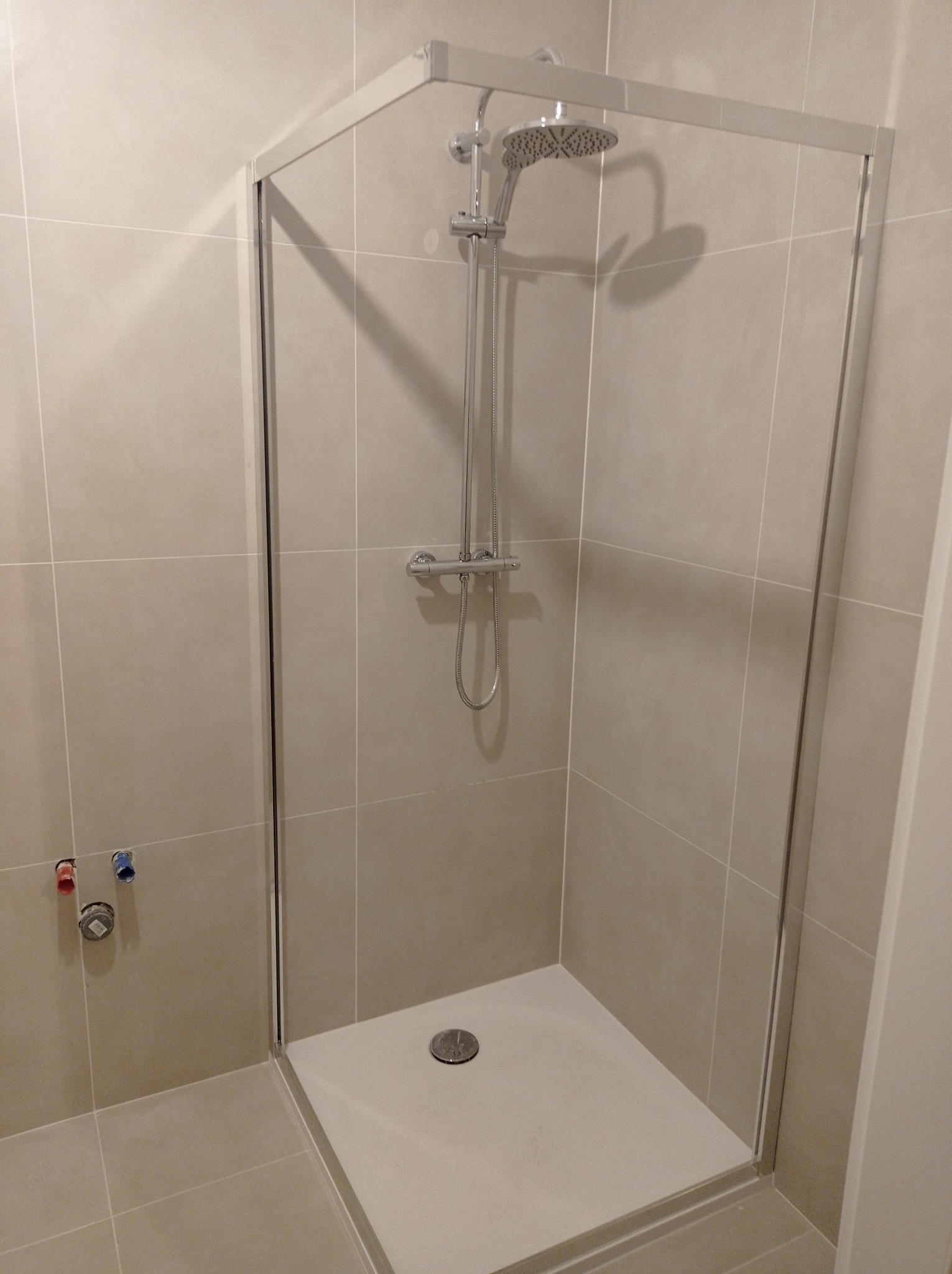 Kabina prysznicowa z brodzikiem i deszczownicą, wykończona beżowymi płytkami. Widoczne zawory wody. Minimalistyczny design, jasne wnętrze.