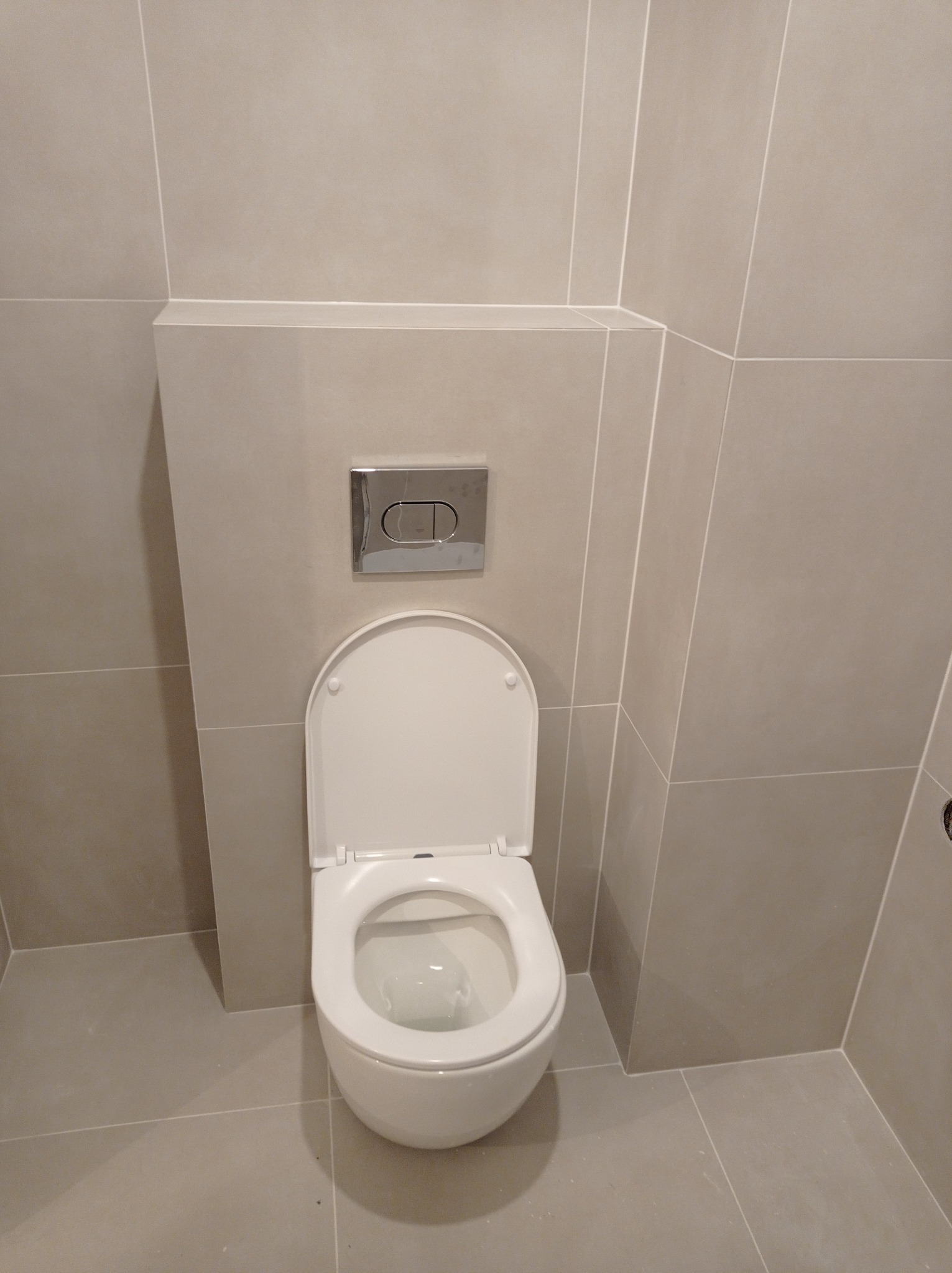 Beżowa, nowoczesna toaleta podwieszana z chromowanym przyciskiem spłukującym, obudowana płytkami w łazience. Minimalistyczny design, jasne wnętrze.