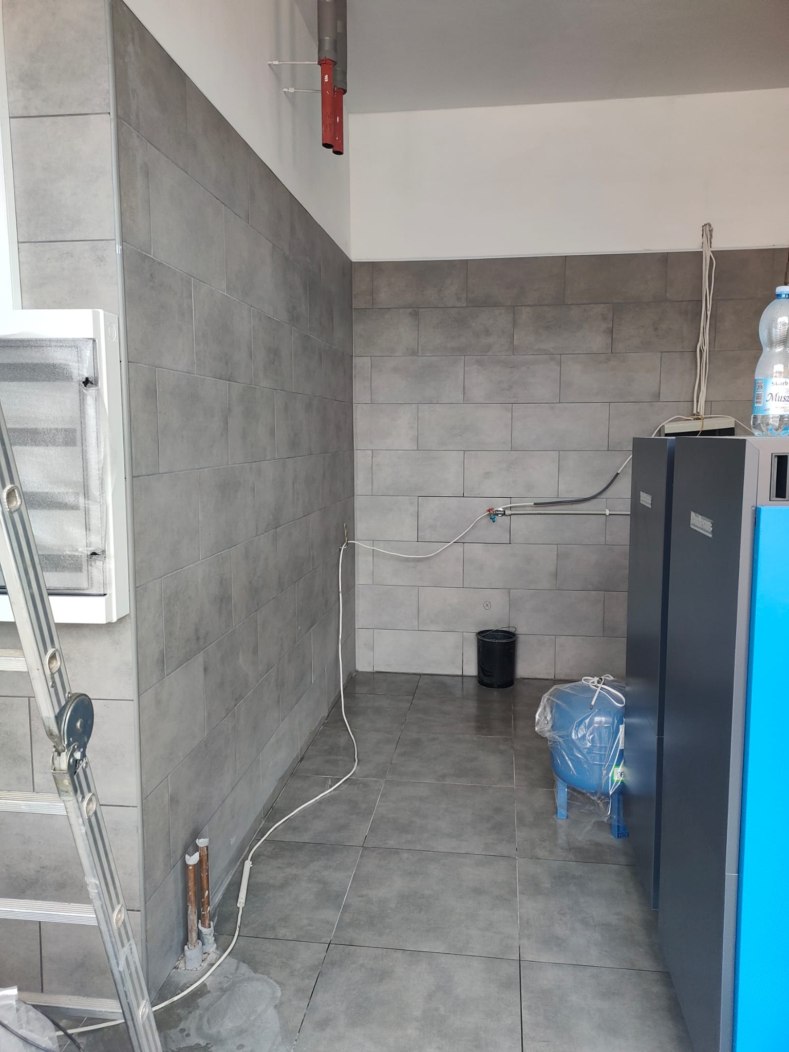 Szare, prostokątne płytki ścienne i podłogowe w pomieszczeniu z instalacją grzewczą. Widoczna drabina, rury i zbiornik na wodę. Minimalistyczny design.