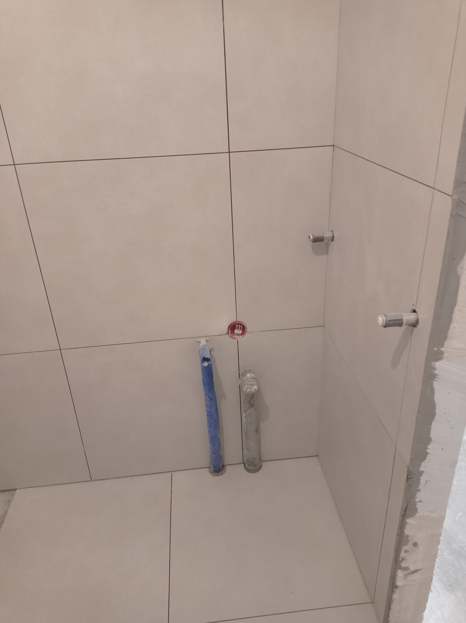 Beżowe płytki ścienne i podłogowe w trakcie montażu w łazience, widoczne rury i instalacja wodna, surowy beton na krawędzi ściany.