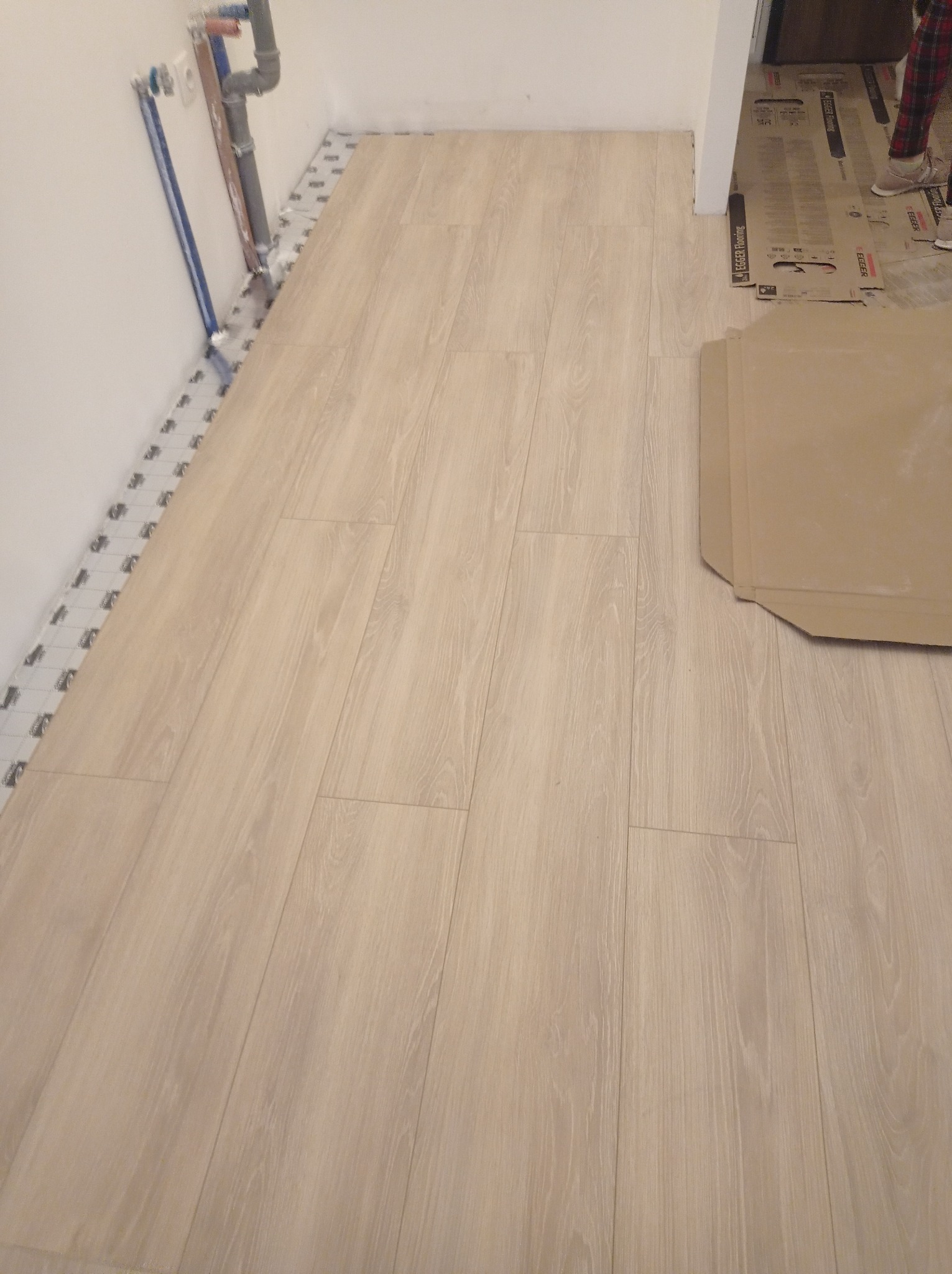 Jasne panele podłogowe ułożone wzdłuż ściany z widocznymi rurami. Część podłogi zabezpieczona kartonem. Widoczna mata pod panele.