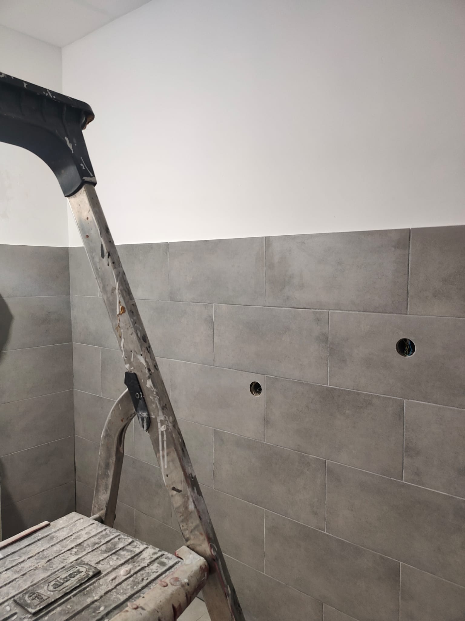 Ściana w szarych, podłużnych płytkach, z widocznymi otworami na gniazdka elektryczne. Obok stoi drabina malarska, co sugeruje etap prac wykończeniowych.