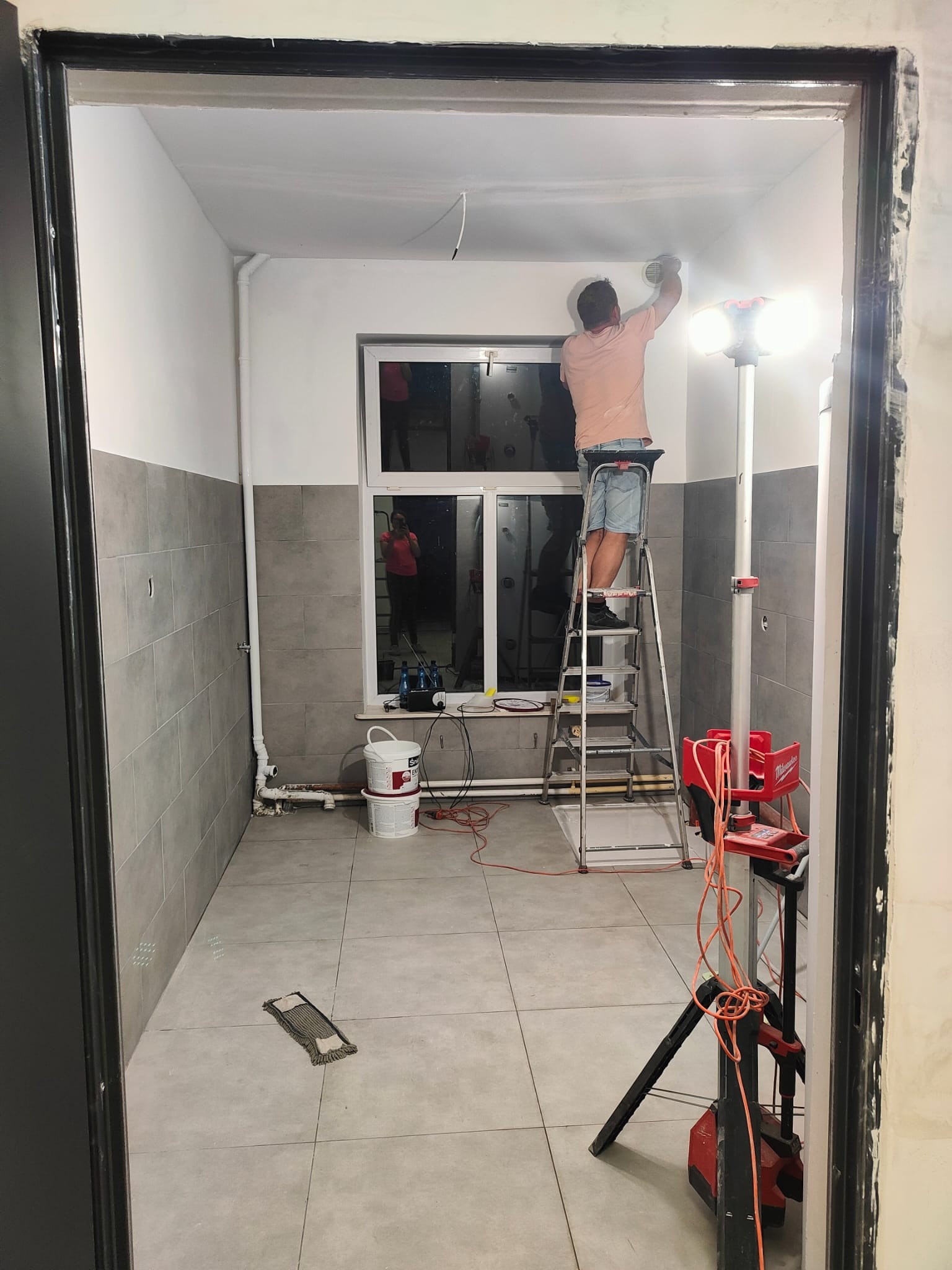 Remont łazienki: mężczyzna na drabinie szlifuje sufit, widoczne płytki na ścianach do połowy wysokości, lampa budowlana i narzędzia. W oknie odbija się osoba.