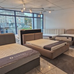 Luxwood Atelier - Salon z ekspozycją łóżek tapicerowanych z materacami. Widoczne różne modele i kolory. Za oknem budynek miejski. Oświetlenie sufitowe punktowe.