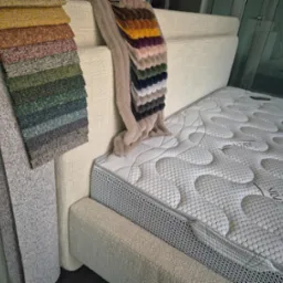 Łóżko tapicerowane z materacem i próbkami tkanin obiciowych w różnych kolorach, prezentowane w Gdyni. Wybór materiałów do mebli na wymiar.