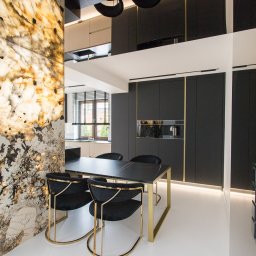 Luxwood Atelier - Elegancka jadalnia z czarnym stołem i krzesłami z złotymi nogami. W tle zabudowa kuchenna w ciemnych kolorach z złotymi akcentami i podświetlana ściana z kamienia.