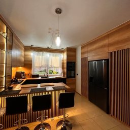 Luxwood Atelier - Nowoczesna kuchnia z drewnianymi lamelami, czarnym blatem i chromowanymi hokerami. Oświetlenie punktowe i okno z firanką dodają przytulności.