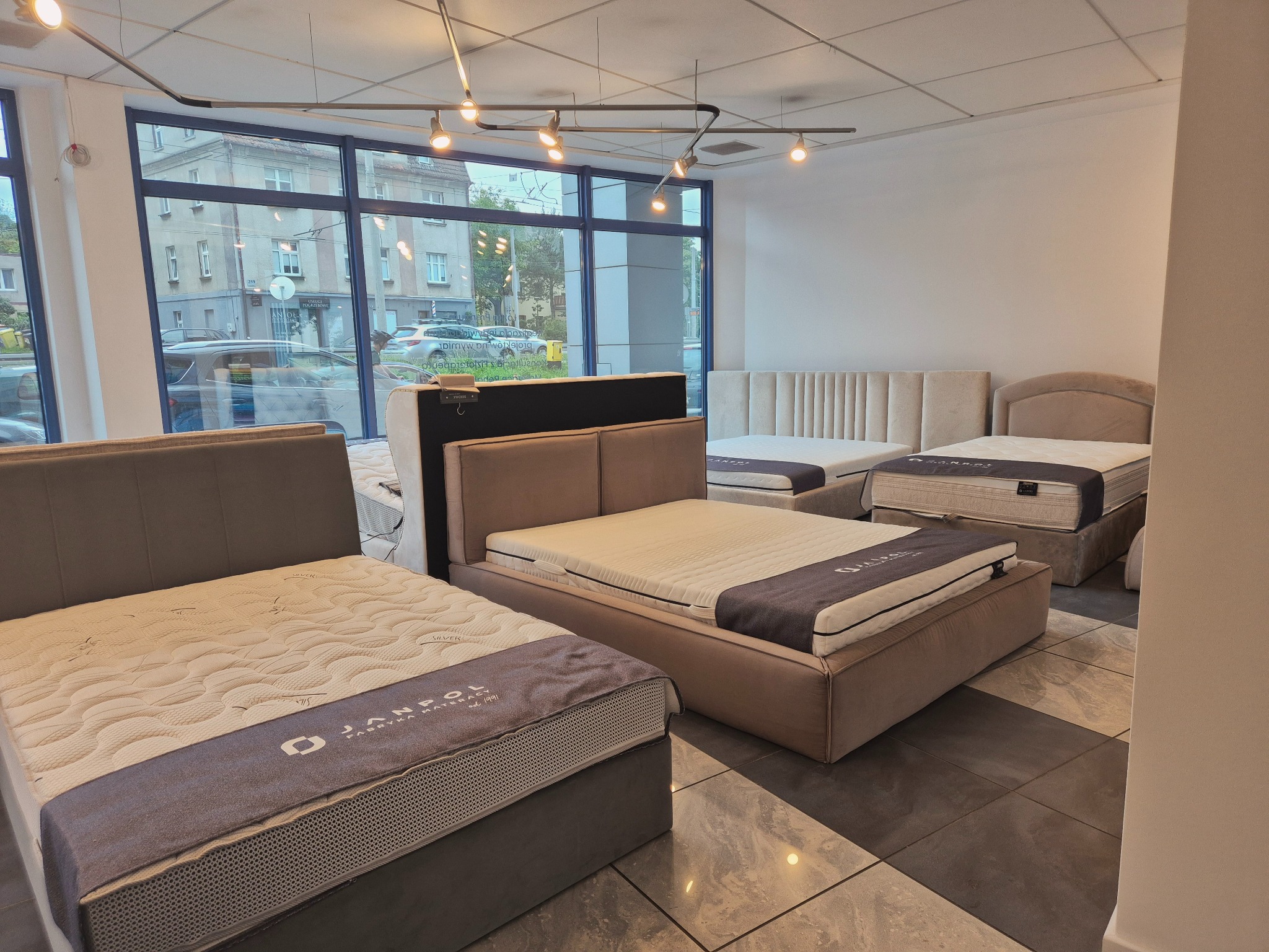 Salon z ekspozycją łóżek tapicerowanych z materacami. Widoczne różne modele i kolory. Za oknem budynek miejski. Oświetlenie sufitowe punktowe.