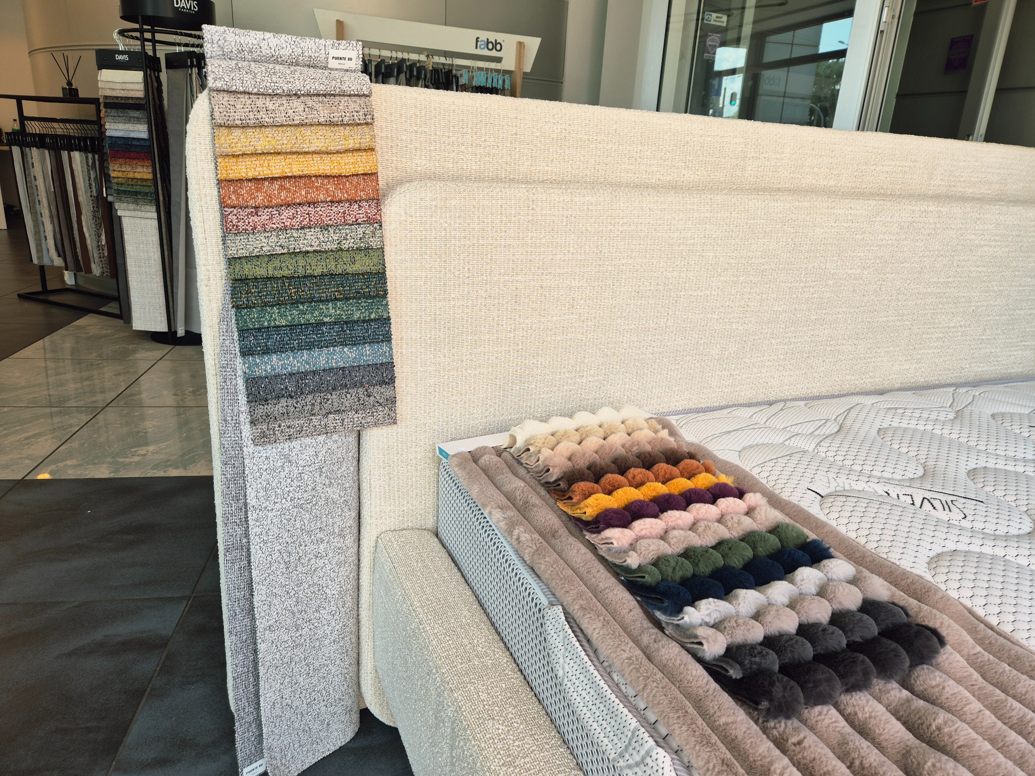 Prezentacja próbek tkanin obiciowych w różnych kolorach i fakturach na tle tapicerowanego zagłówka łóżka. Wzorniki materiałów ekspozycja w salonie meblowym.