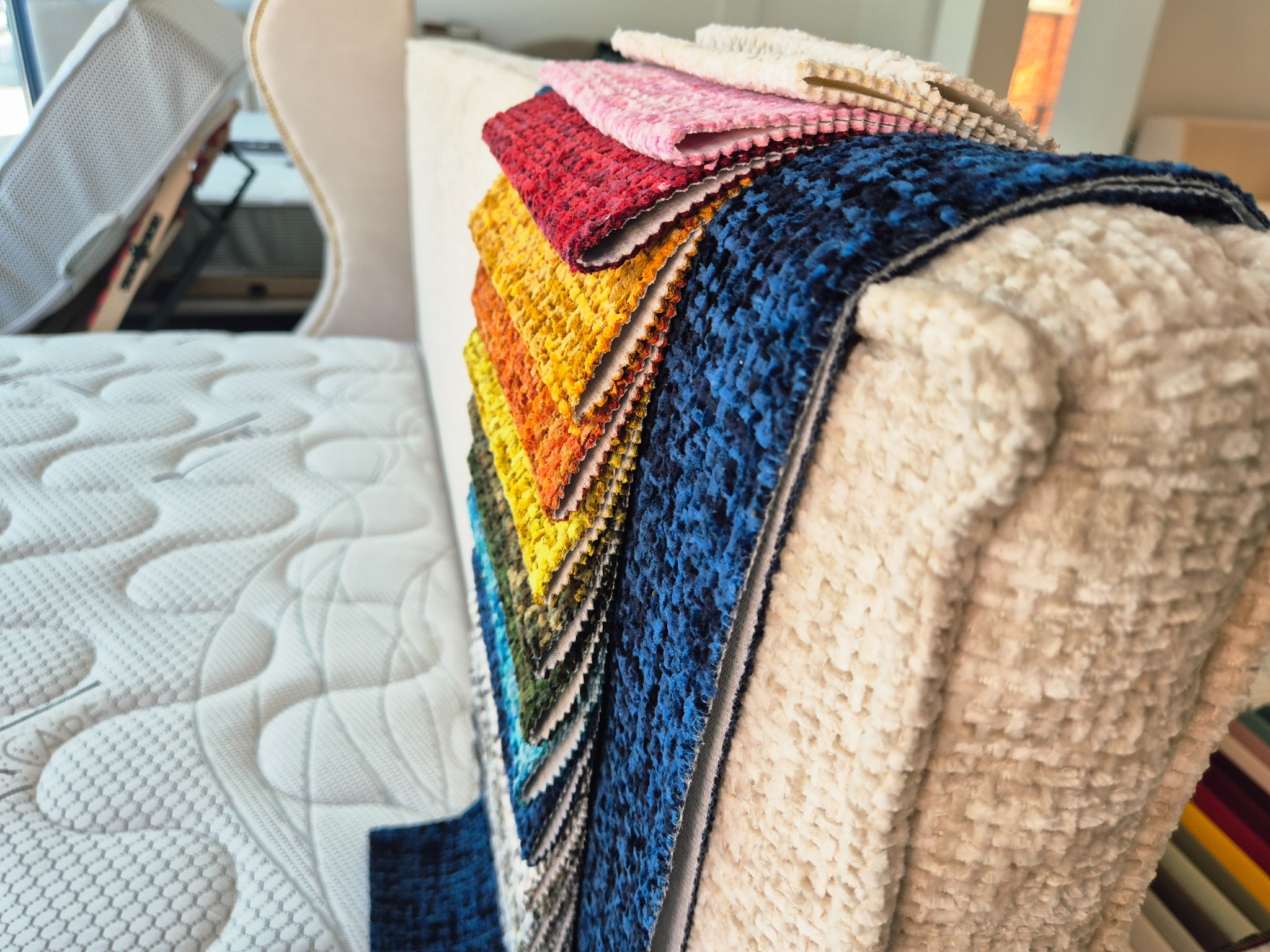 Próbniki kolorów tapicerki ułożone na zagłówku łóżka z pikowanym materacem w tle. Paleta barw od niebieskiego po różowy, tekstura pluszowa.