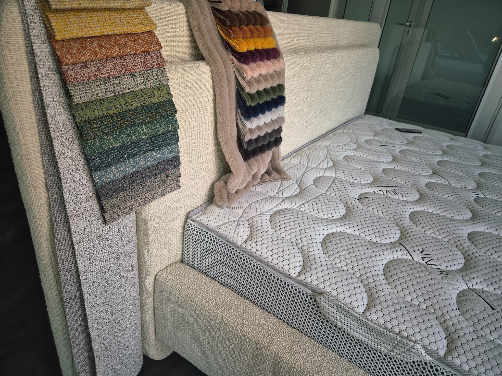 Łóżko tapicerowane z materacem i próbkami tkanin obiciowych w różnych kolorach, prezentowane w Gdyni. Wybór materiałów do mebli na wymiar.