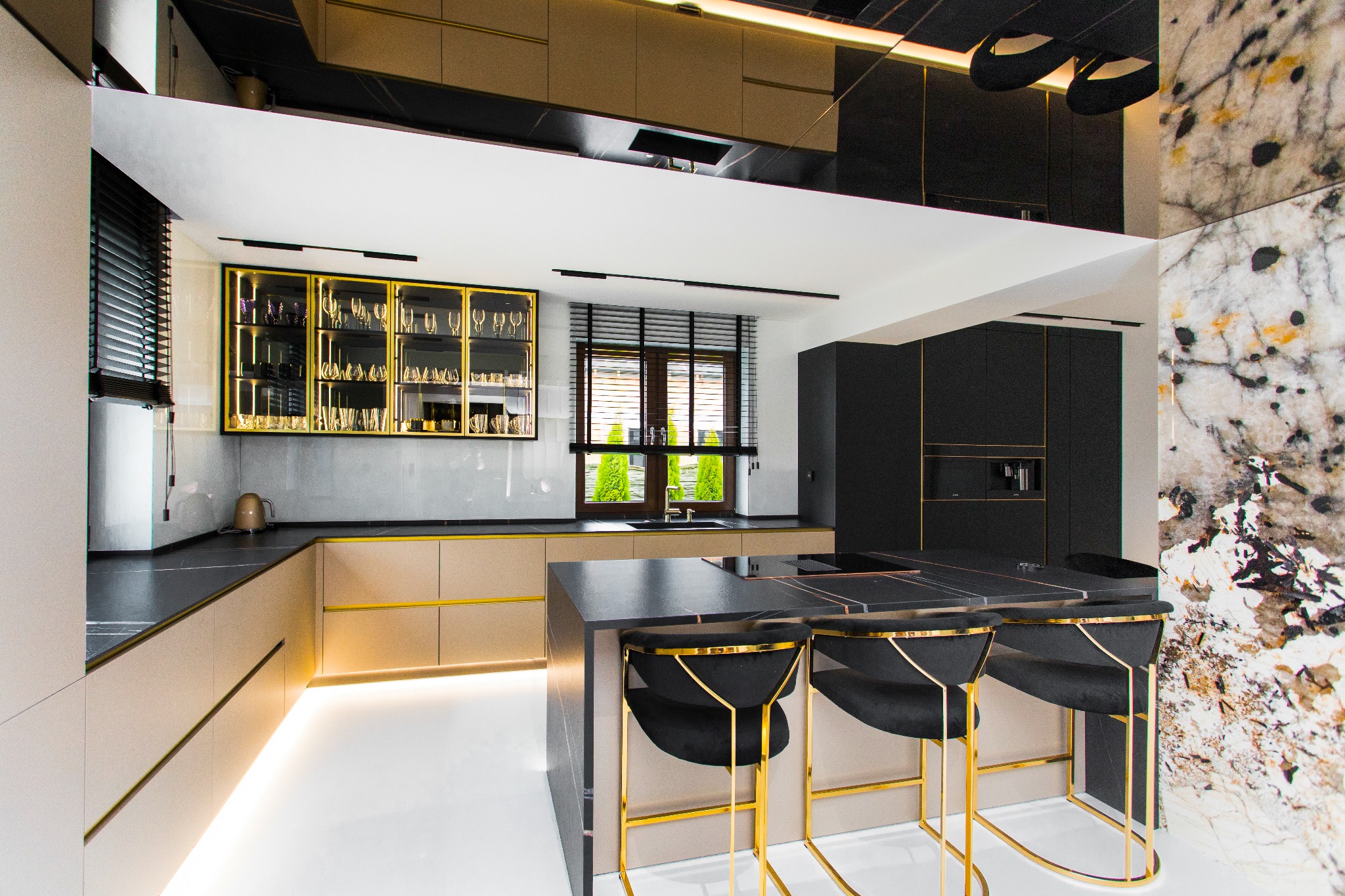Nowoczesna kuchnia: beżowe szafki z złotymi akcentami, czarny blat i wyspa z hokerami. Marmurowa ściana i oświetlenie LED. Minimalistyczny design z luksusowymi detalami.