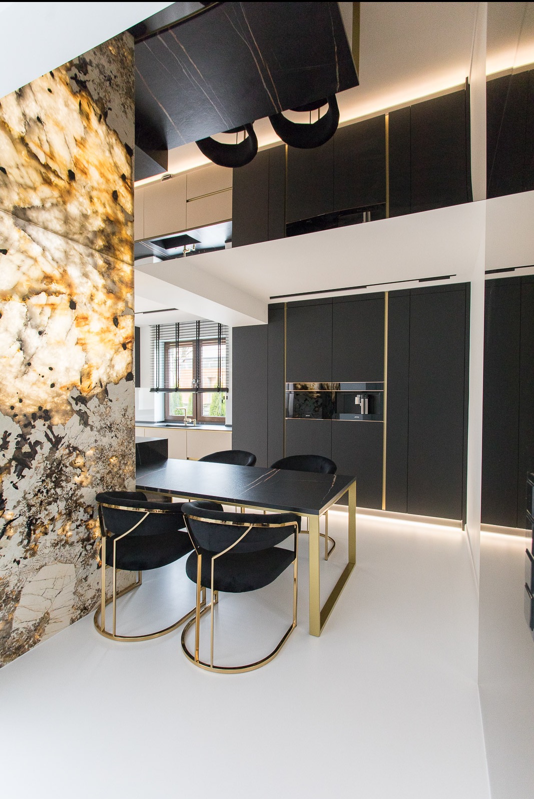 Elegancka jadalnia z czarnym stołem i krzesłami z złotymi nogami. W tle zabudowa kuchenna w ciemnych kolorach z złotymi akcentami i podświetlana ściana z kamienia.