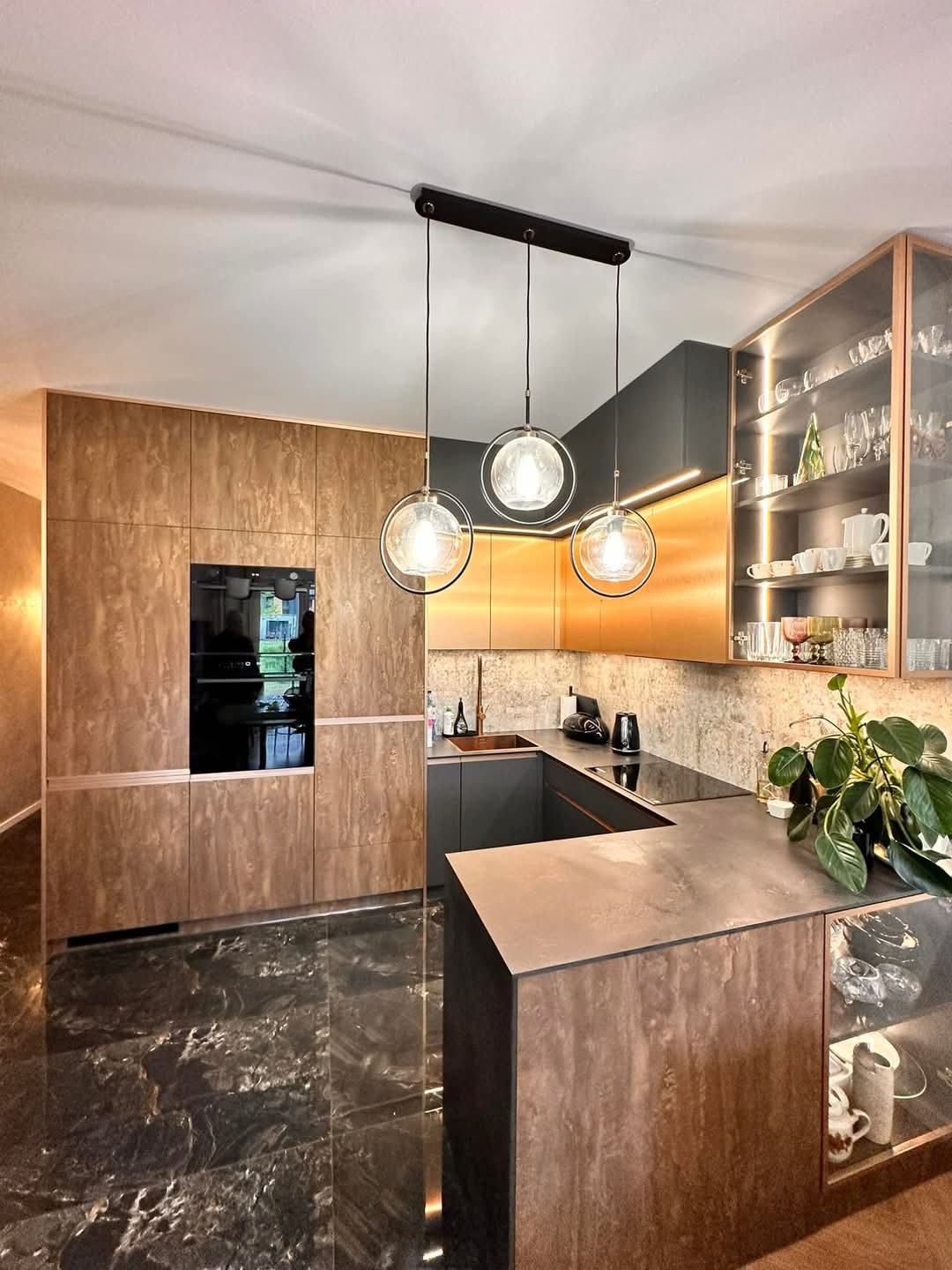 Nowoczesna kuchnia z marmurową podłogą, drewnianymi frontami, szarymi elementami i trzema okrągłymi lampami wiszącymi. Szkło i oświetlenie w szafkach.