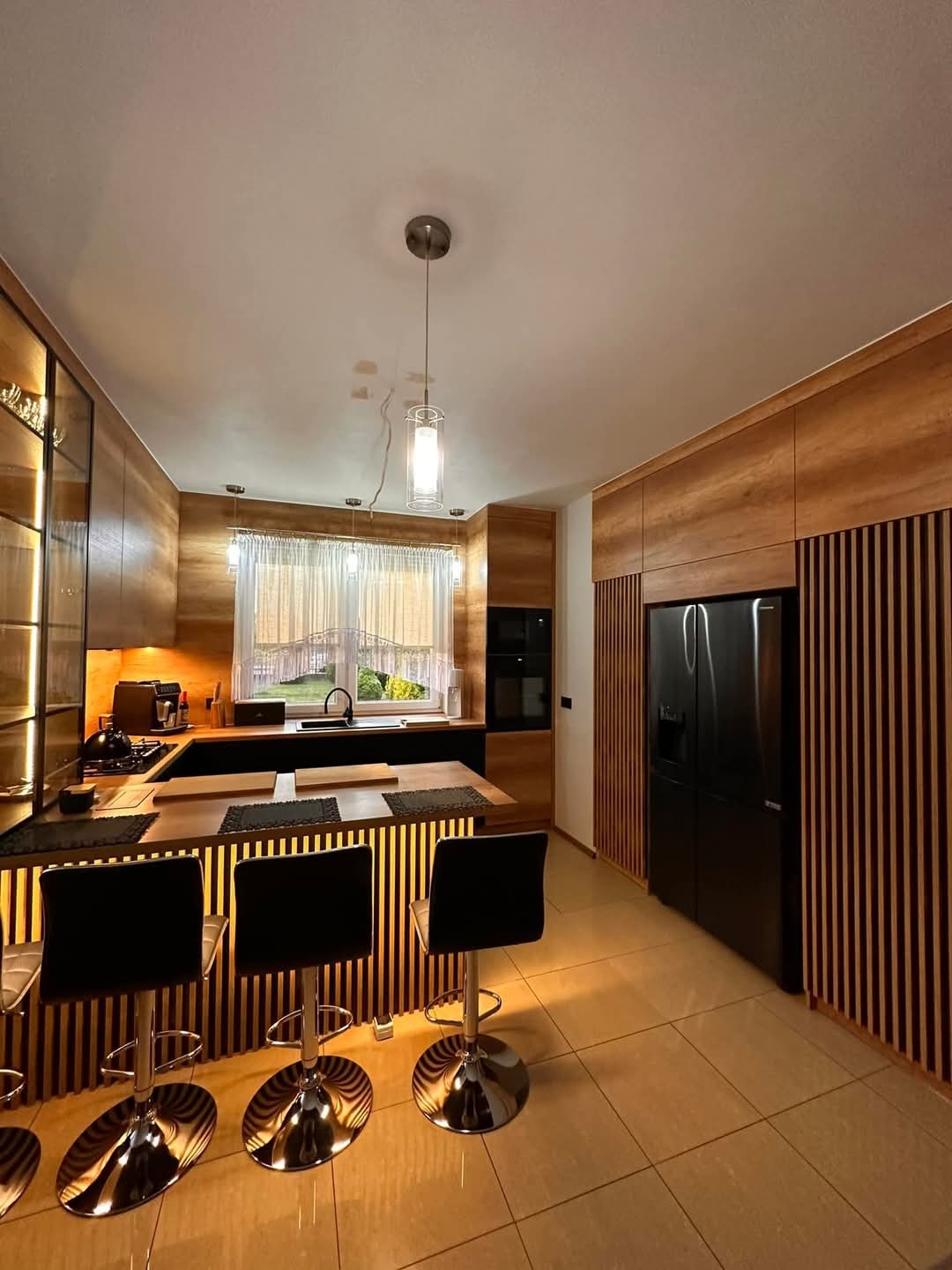 Nowoczesna kuchnia z drewnianymi lamelami, czarnym blatem i chromowanymi hokerami. Oświetlenie punktowe i okno z firanką dodają przytulności.
