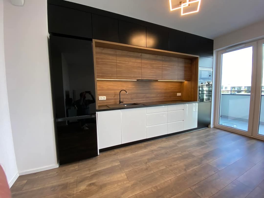 Nowoczesna kuchnia: czarne szafki górne, drewniane wnęki, białe szafki dolne z błyszczącymi frontami. Widoczne okno balkonowe i drewniana podłoga.