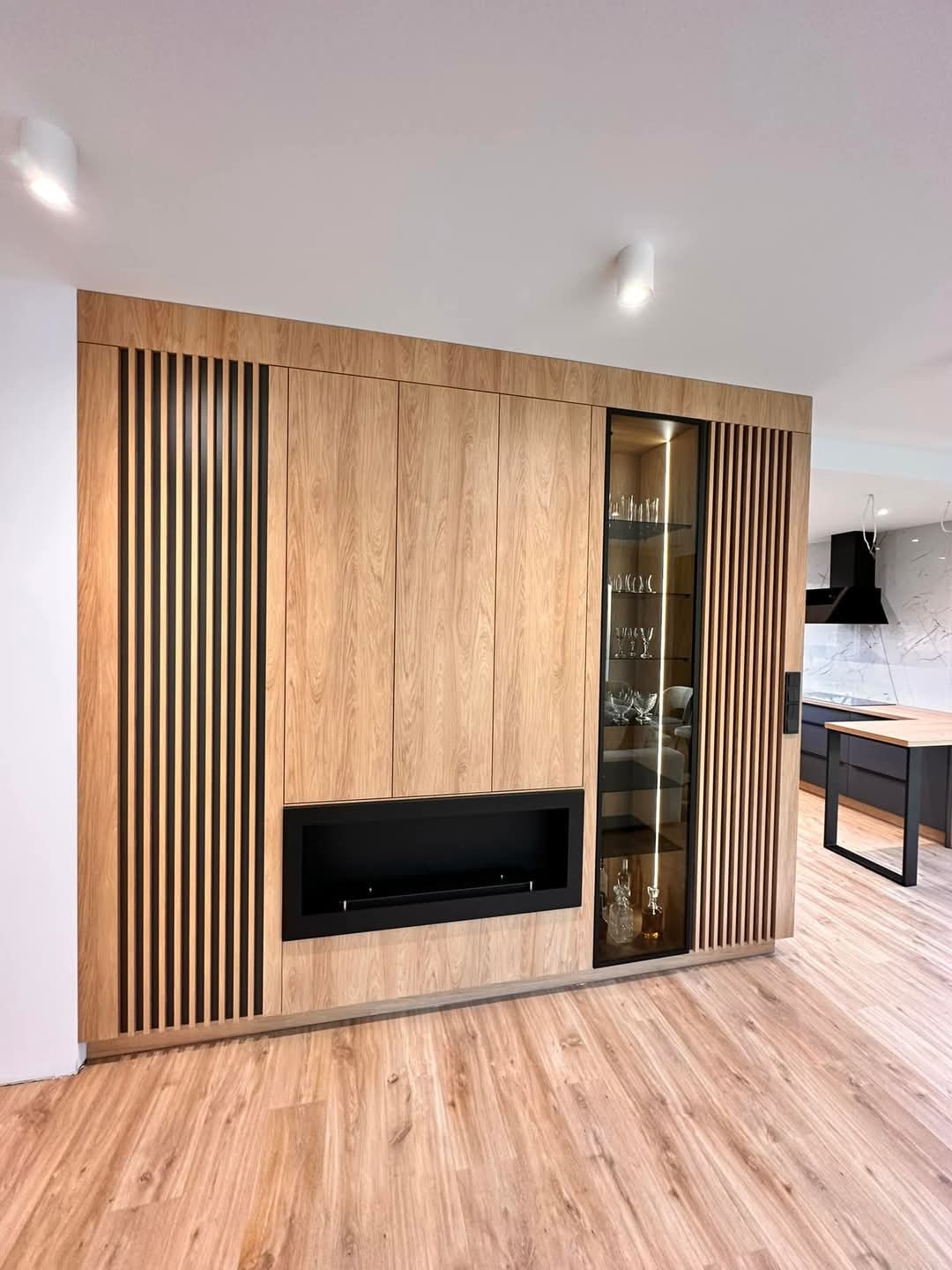 Drewniana zabudowa ściany z szafkami, kominkiem i witryną z podświetleniem. Na podłodze panele imitujące drewno. W tle widoczna kuchnia z czarnymi szafkami.