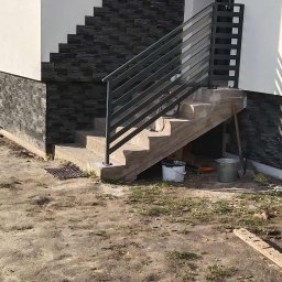Lidia Tadych - Schody zewnętrzne z szarą balustradą i okładziną elewacyjną imitującą kamień w Kleszczynie. Widoczne narzędzia i wiaderka, sugerujące prace wykończeniowe.