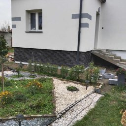 Lidia Tadych - Wykończona elewacja domu z szarym pasem i kamienną podmurówką. Widoczny fragment ogrodu z kwiatami i ścieżką z kamyków. Ujęcie z niskiej perspektywy.