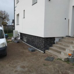 Lidia Tadych - Wykończona elewacja domu z dekoracyjnym kamieniem na cokole. Widoczne schody wejściowe i fragment instalacji klimatyzacji. Biała elewacja, szara kostka.