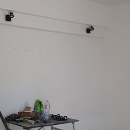 Lidia Tadych - Biała ściana po malowaniu z zamontowanym oświetleniem punktowym na szynoprzewodzie. Na pierwszym planie rusztowanie z narzędziami malarskimi.