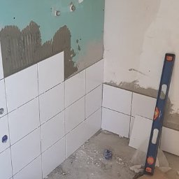 Lidia Tadych - Białe płytki ułożone do połowy ściany w rogu łazienki, widoczny poziomica i resztki zaprawy. Etap prac wykończeniowych w trakcie układania glazury.
