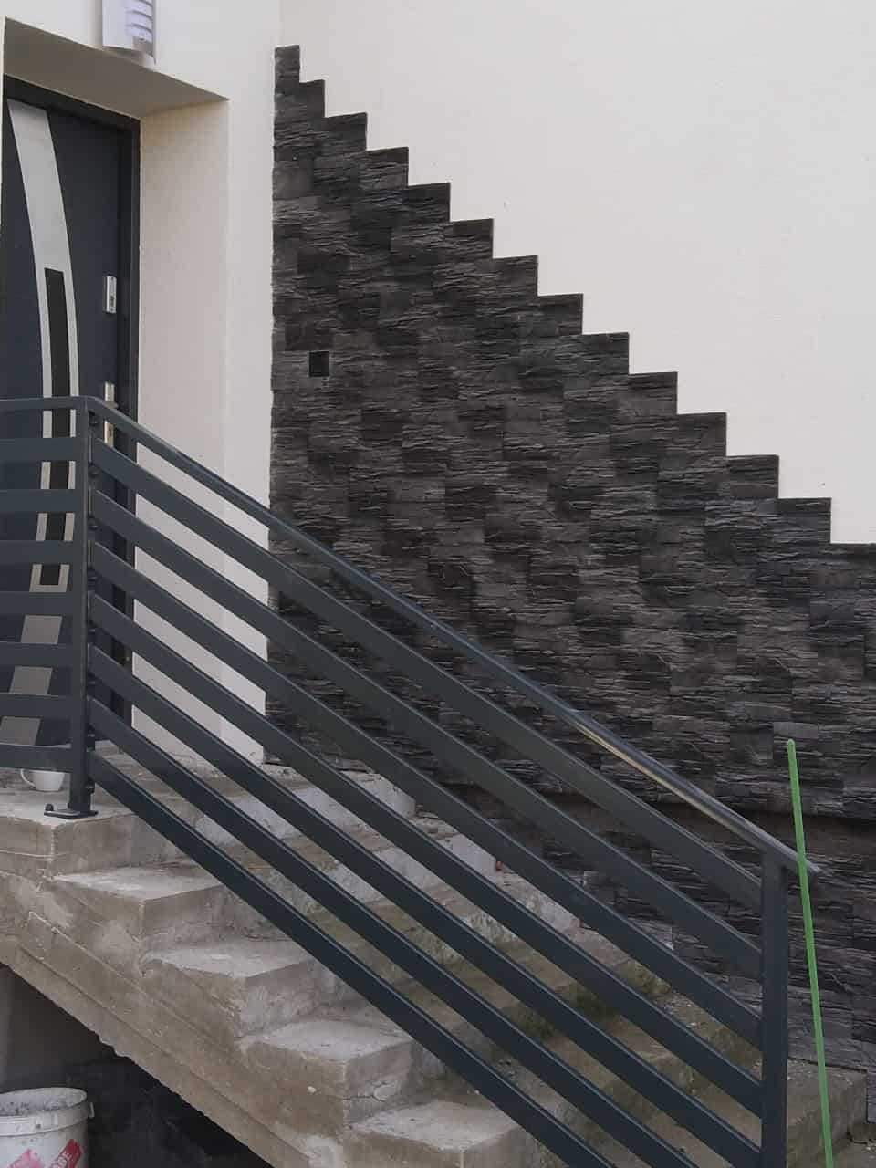 Schody wejściowe z szarą balustradą i dekoracyjną, ciemną okładziną elewacyjną ułożoną schodkowo. Nowoczesne wykończenie zewnętrzne budynku.