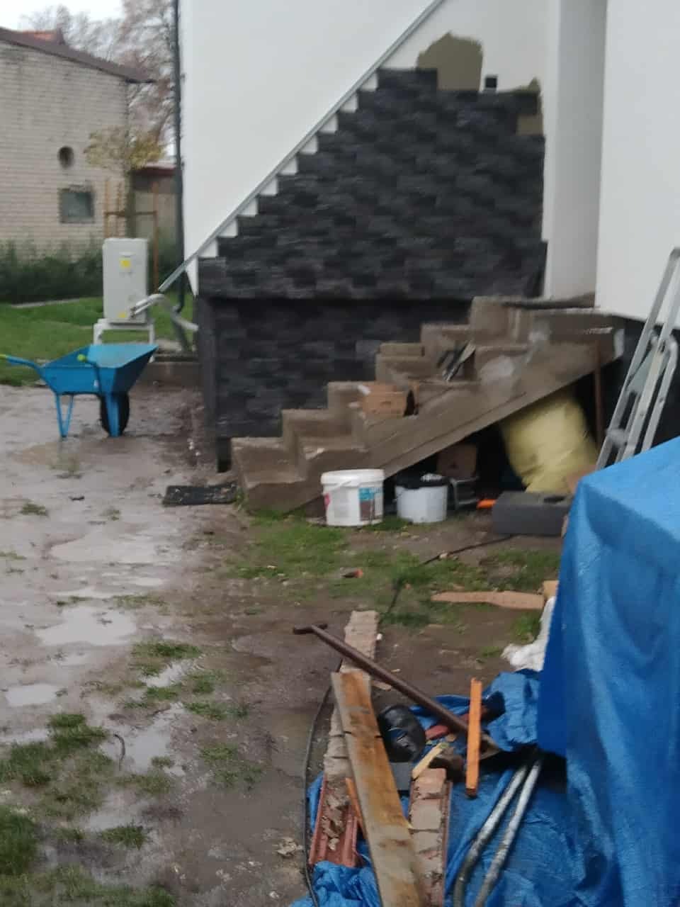 Montaż elewacji z kamienia dekoracyjnego na schodach zewnętrznych budynku. Widoczne narzędzia i materiały budowlane. Prace w toku, etap montażu.