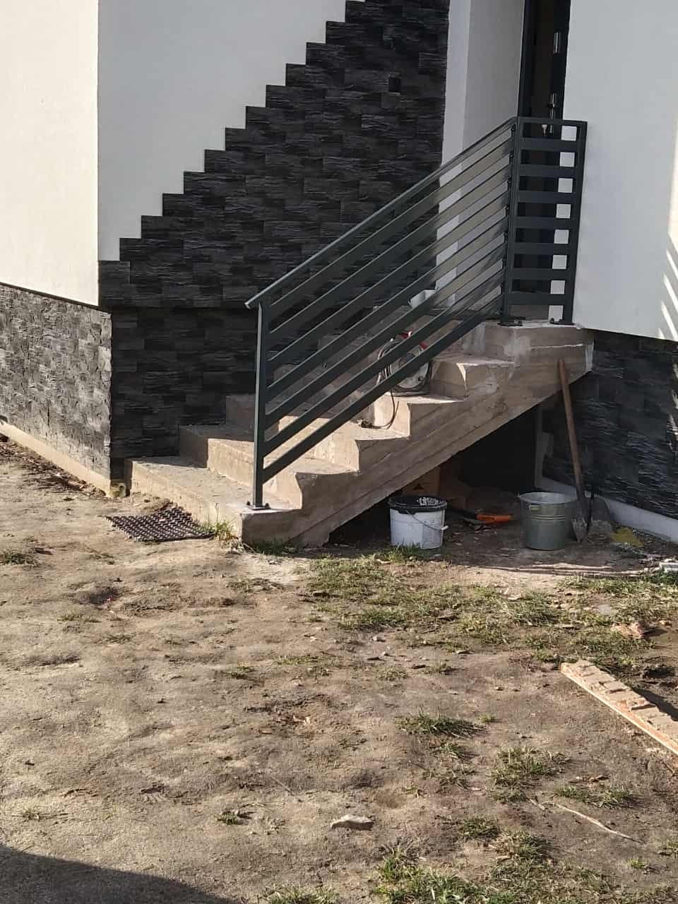Schody zewnętrzne z szarą balustradą i okładziną elewacyjną imitującą kamień w Kleszczynie. Widoczne narzędzia i wiaderka, sugerujące prace wykończeniowe.