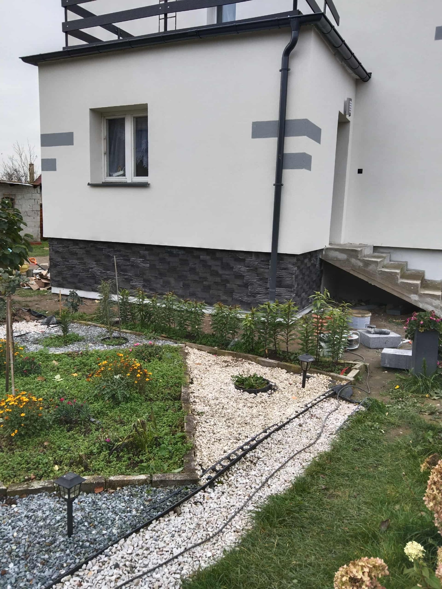 Wykończona elewacja domu z szarym pasem i kamienną podmurówką. Widoczny fragment ogrodu z kwiatami i ścieżką z kamyków. Ujęcie z niskiej perspektywy.