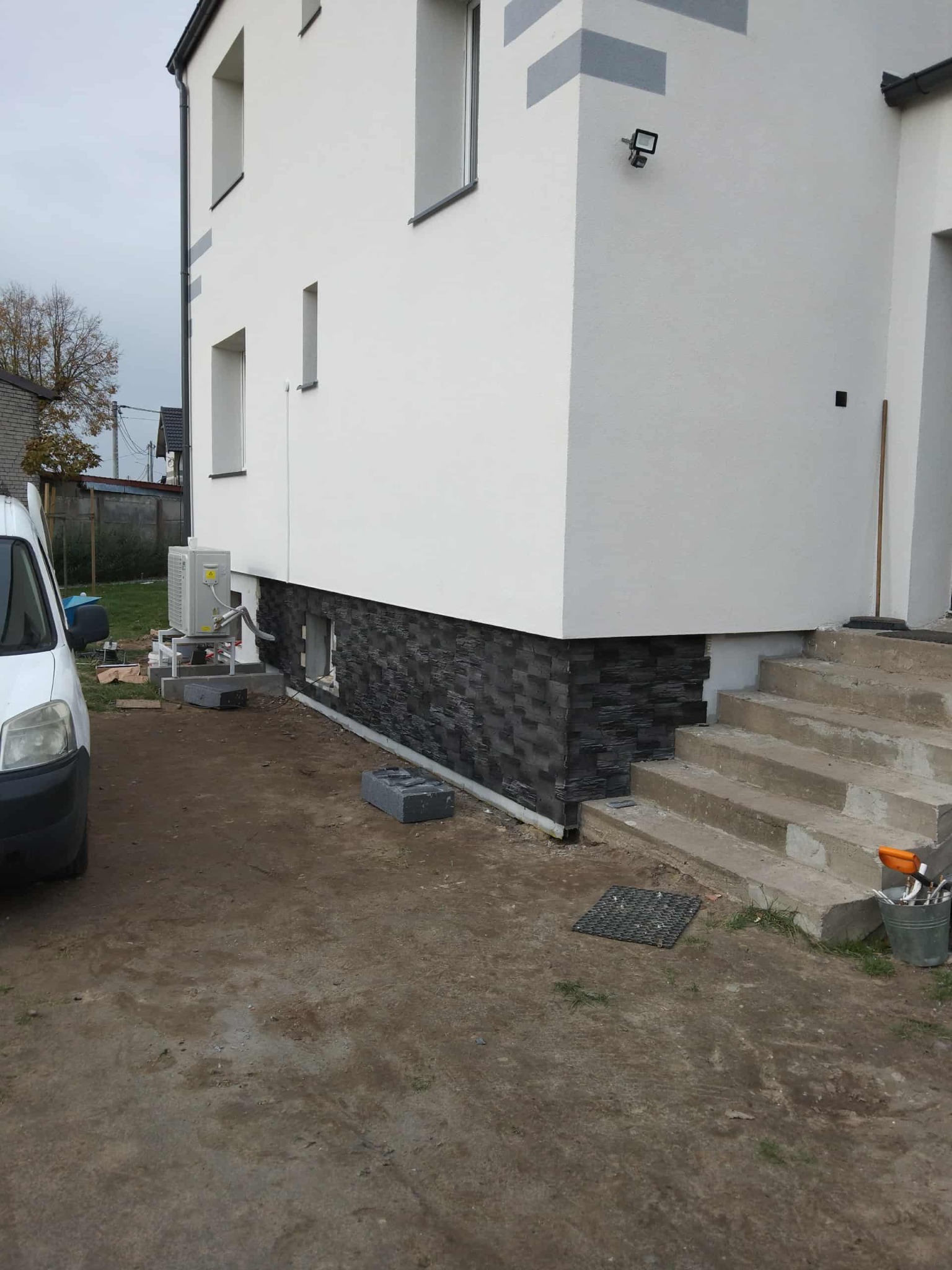 Wykończona elewacja domu z dekoracyjnym kamieniem na cokole. Widoczne schody wejściowe i fragment instalacji klimatyzacji. Biała elewacja, szara kostka.
