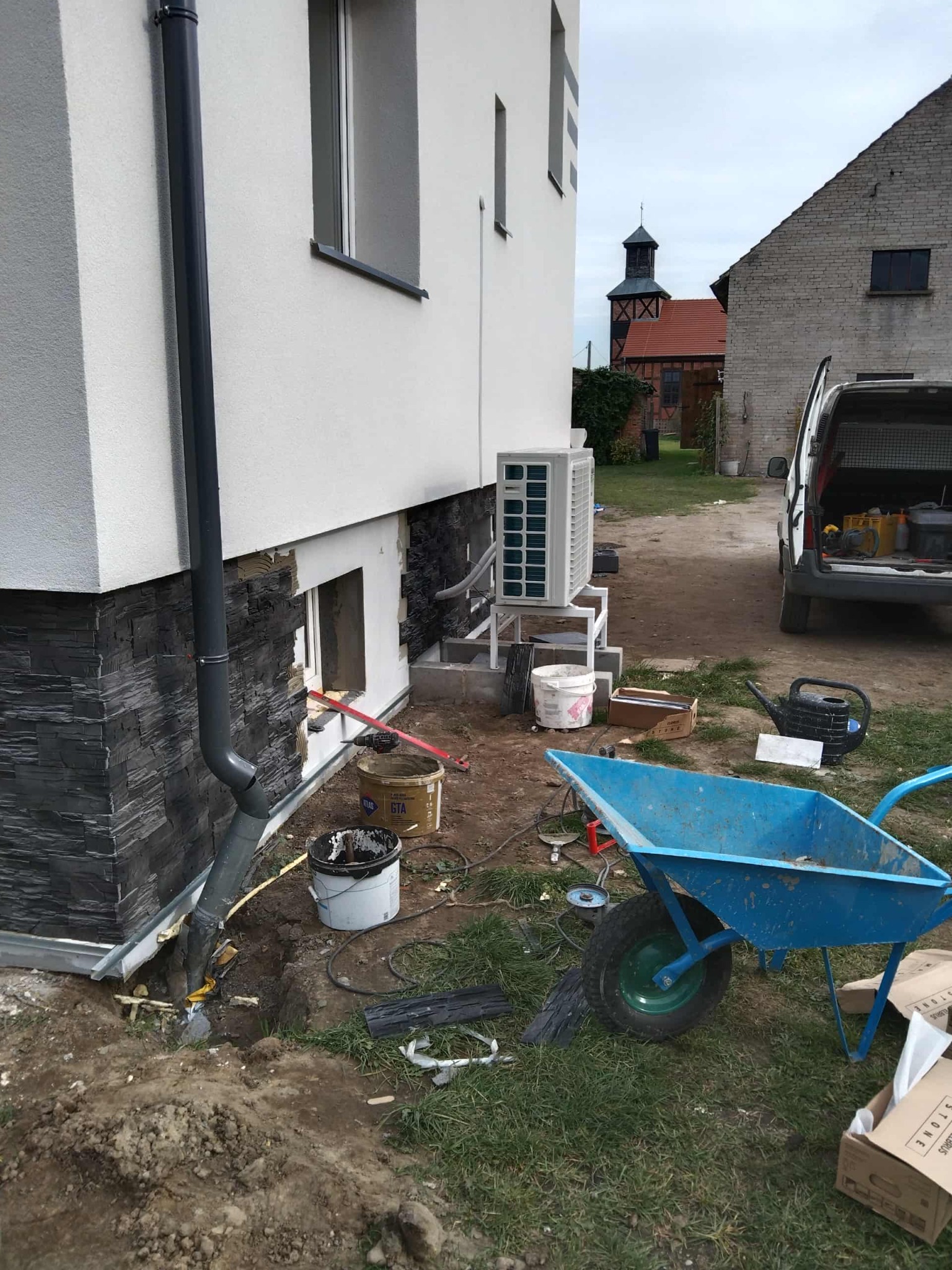 Fragment elewacji budynku z wykończeniem z kamienia dekoracyjnego, widoczne narzędzia i materiały budowlane, w tle budynek gospodarczy i kościół.