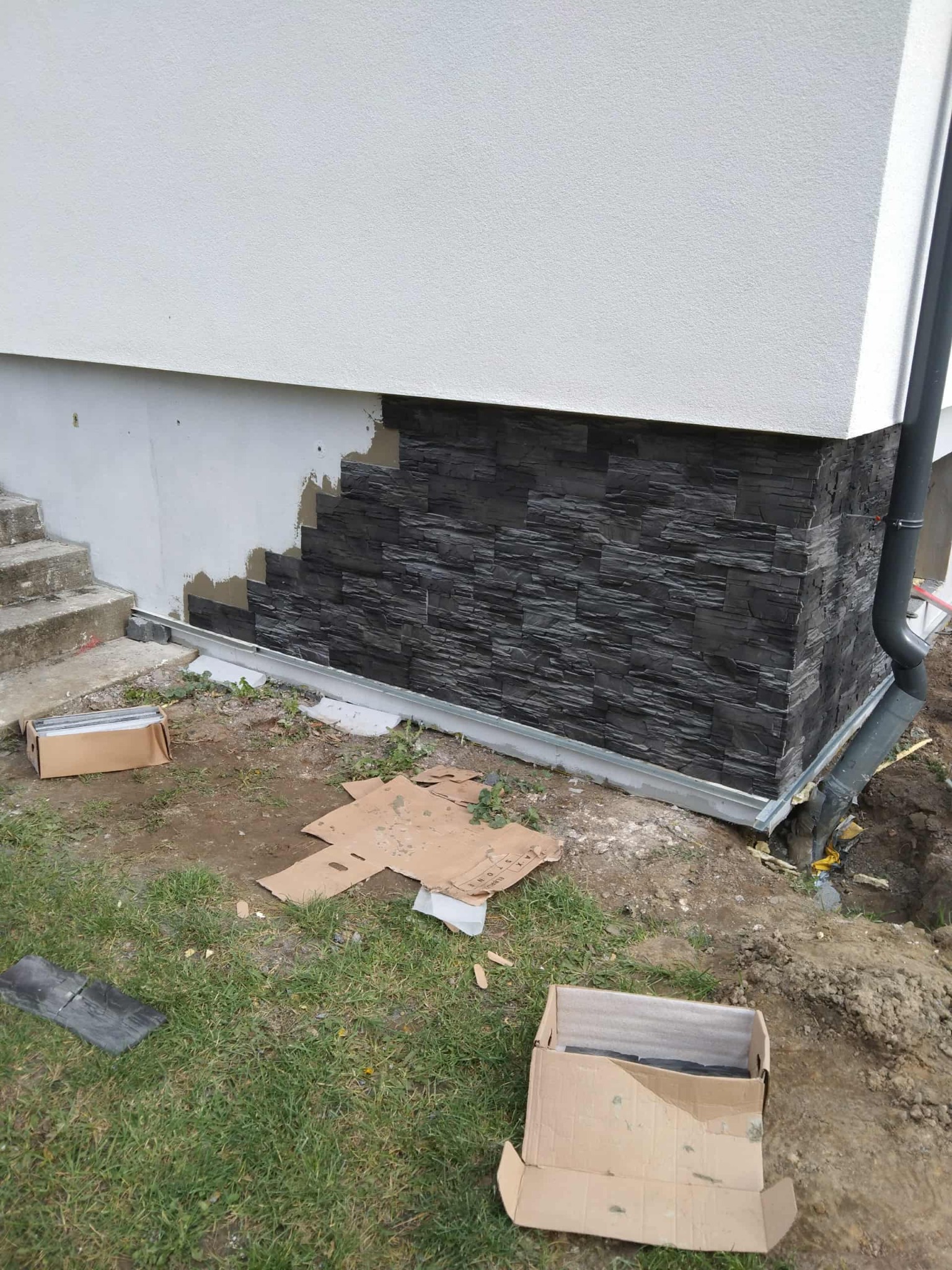 Fragment elewacji budynku z częściowo ułożoną, ciemną okładziną kamienną. Widoczne kartony z materiałem i fragment trawnika. Ujęcie z bliska, w trakcie prac.