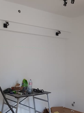 Biała ściana po malowaniu z zamontowanym oświetleniem punktowym na szynoprzewodzie. Na pierwszym planie rusztowanie z narzędziami malarskimi.