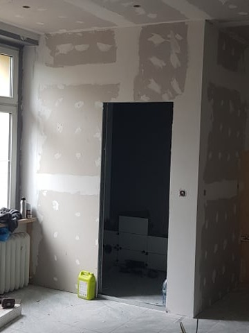 Wnętrze w trakcie prac wykończeniowych: ściany z płyt gipsowo-kartonowych, częściowo zaszpachlowane, widoczne wejście do pomieszczenia z ułożonymi płytkami.