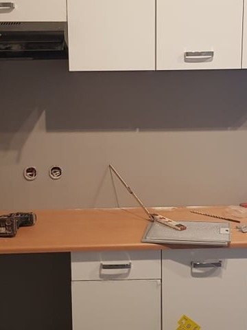 Kuchnia w trakcie montażu: białe szafki, drewniany blat, narzędzia (wiertarka, poziomica) i puszki elektryczne na szarej ścianie. Widok na etap prac.