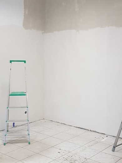Pokój w trakcie malowania: ściany częściowo pokryte farbą, drabina aluminiowa z zielonymi stopniami, płytki podłogowe. Widoczny etap prac wykończeniowych.