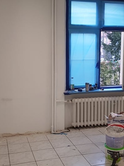 Pokój w trakcie malowania: ściana w kolorze złamanej bieli, okno zabezpieczone folią, grzejnik i wiaderka z farbą. Widoczne przygotowania do malowania.