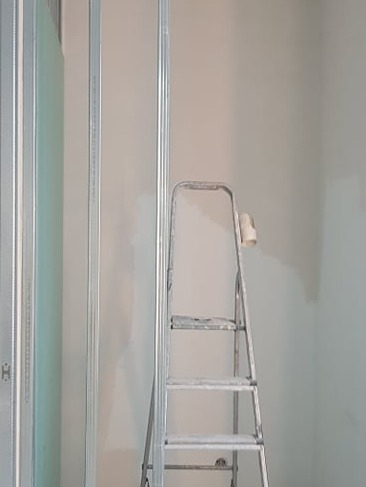 Stelaż z płyt gipsowo-kartonowych przy ścianie, drabina malarska z wałkiem, przygotowanie do malowania ścian w jasnym wnętrzu. Prace wykończeniowe w trakcie.