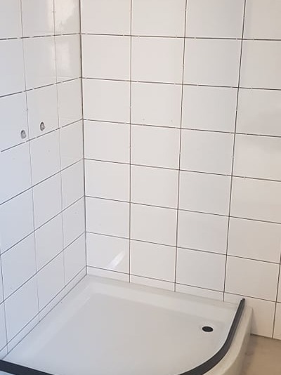 Biały brodzik z czarnym obramowaniem w rogu łazienki wyłożonej białymi, kwadratowymi płytkami. Widoczne otwory na armaturę.