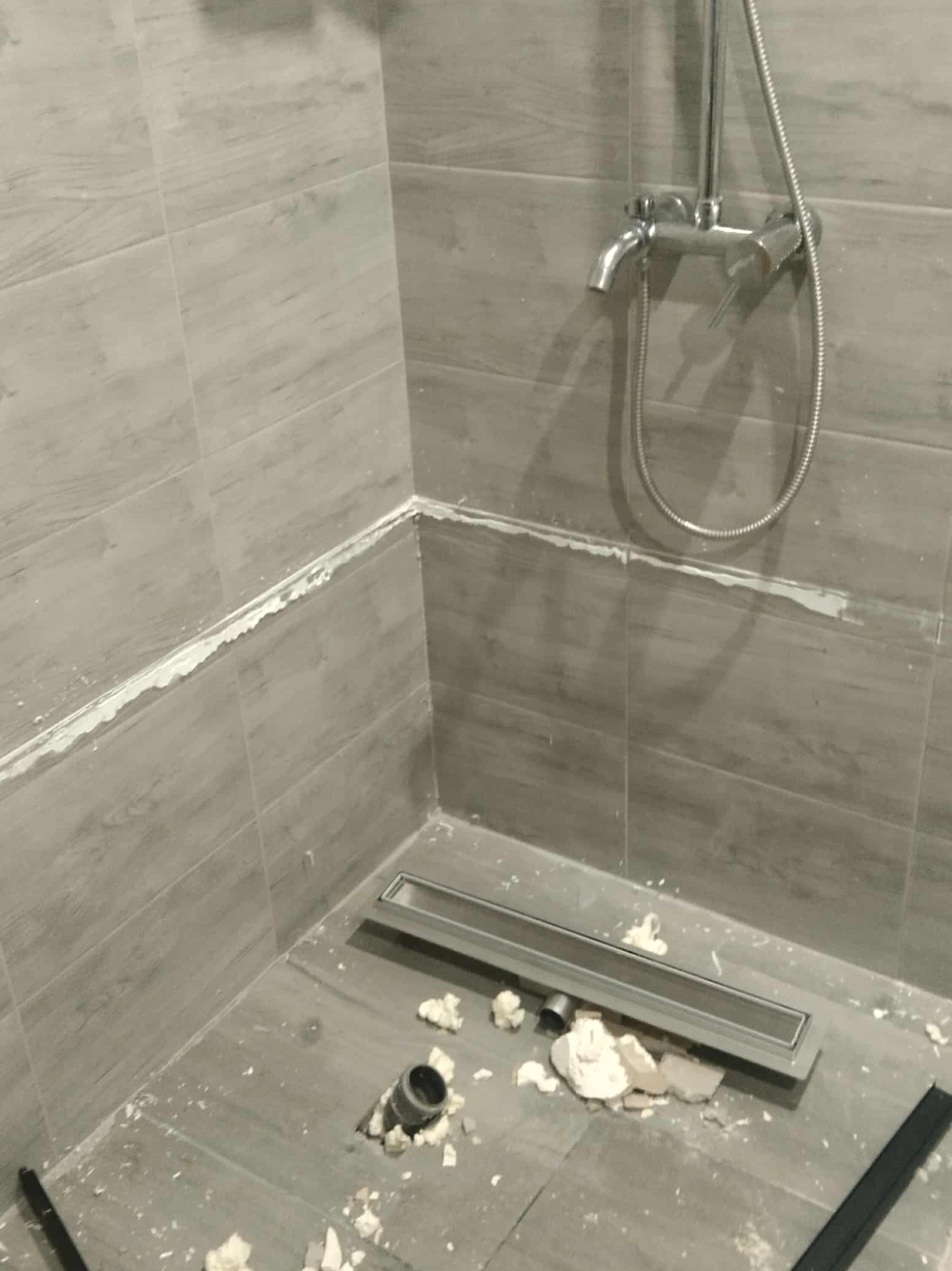 Szara łazienka w trakcie prac: ułożone płytki imitujące drewno, zamontowany odpływ liniowy, widoczna armatura prysznicowa i ślady po fugowaniu.